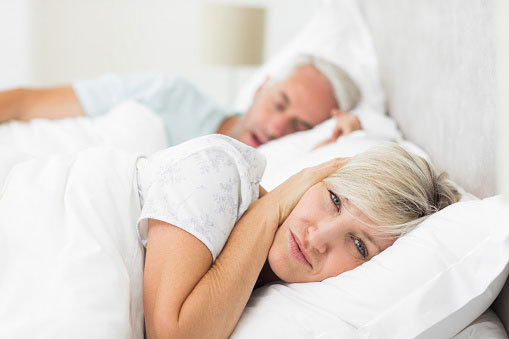 Couple in bed with husband snoring and wife covering her ears.