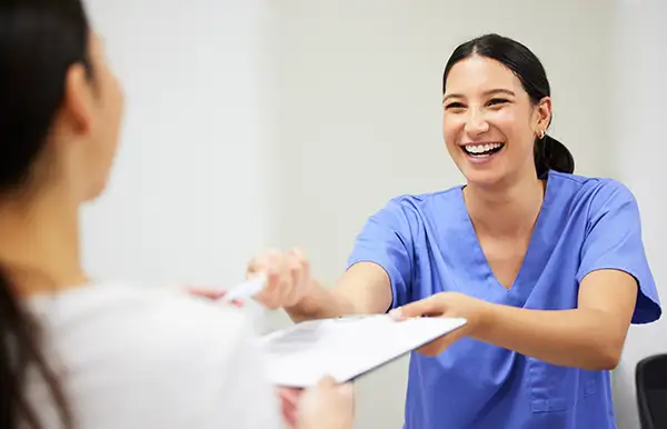 Smiling dental office receptionist in scrubs handing new patient forms to a patient at the reception desk.